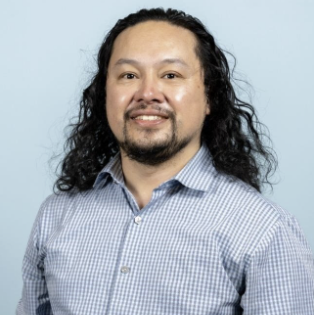 Headshot of a lightly tan skinned Filipino man with long black curly hair wearing a blue dress shirt smiling at the camera.